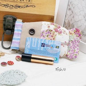 May include: A handmade, fish-shaped fabric wallet with floral and striped patterns. The wallet features a button closure and holds a blue and white card. A gold and black tube of lipstick and decorative buttons are also present.