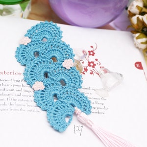 May include: A blue crocheted bookmark with pink flowers and a pink tassel. The bookmark is shaped like a leaf and has a scalloped edge.