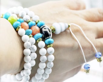May include: A white beaded bracelet with turquoise, orange, and green accents. The bracelet has a white shell charm and a tassel with blue crystals.