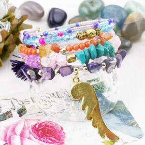 May include: A stack of colorful bracelets with beads, stones, and a gold wing charm. The bracelets are made with various materials, including turquoise, amethyst, and coral.