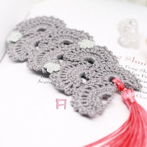 May include: Gray crocheted bookmark with a pink tassel and three white crocheted flowers. The bookmark is on a white page with the text "Feng Shui" visible.