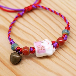Könnte beinhalten: Ein rotes und violettes Armband aus Schnur mit einem Keramik-Katzenanhänger, roten und orangefarbenen Perlen und einer kleinen Glocke. Der Anhänger trägt das japanische Wort "福" (bedeutet "Glück").