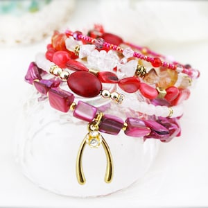 Könnte beinhalten: Ein Stapel bunter Armbänder mit rosa, roten und weißen Perlen, akzentuiert mit goldenen Akzenten und einem goldenen Hufeisen-Anhänger.