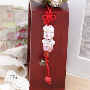 Puede incluir: Un cordón rojo con un nudo decorativo y tres figuras de gato de la suerte de cerámica blanca. Los gatos tienen caras pintadas y detalles florales. Una pequeña campana y cuentas rojas cuelgan de la parte inferior. Fondo de madera rojo oscuro.