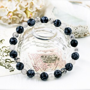Puede incluir: Un brazalete de piedra moteada en blanco y negro con cuentas transparentes. El brazalete está sobre una superficie blanca con un cuenco de vidrio y un trozo de encaje.