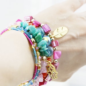 May include: A stack of colorful bracelets with turquoise, pink, and gold beads. The bracelets feature a gold charm with a hamsa hand and a gold feather charm.