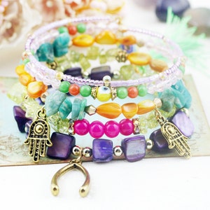 Könnte beinhalten: Mehrfarbiges Perlenarmband-Set mit Charms. Das Set enthält ein lila, pink, grün, orange und gelbes Perlenarmband mit einem goldenen Hamsa-Hand-Anhänger und einem Hufeisen-Anhänger.