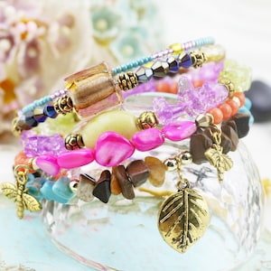 May include: A stack of colorful beaded bracelets with various charms. The bracelets feature pink, brown, and turquoise beads, along with gold-colored dragonfly and leaf charms. The bracelets are stacked on a clear glass dish.