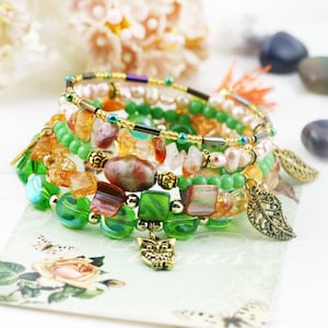 Könnte beinhalten: Ein Set aus sieben Armbändern mit verschiedenen Perlen, darunter grüne, orange und braune. Die Armbänder sind übereinander gestapelt und beinhalten einen kleinen Eulen-Anhänger.