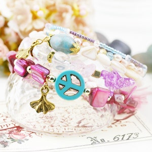 May include: A stack of colorful bracelets with pink, purple, blue, and white beads. The bracelets feature a peace sign charm, a ginkgo leaf charm, and a turquoise stone charm.