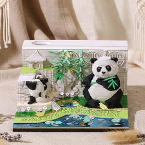 Puede incluir: Diorama de oso panda impreso en 3D con un panda blanco y negro comiendo bambú. La escena incluye un estanque con nenúfares, bambú y una pequeña figura de panda. El diorama está sobre un fondo blanco.