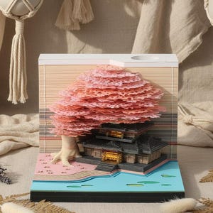 May include: A layered paper art piece depicting a Japanese temple scene with a pink cherry blossom tree. The artwork features a detailed building, a blue pond, and a light-colored background. The base is black.