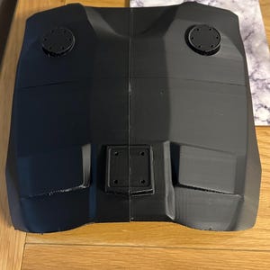 May include: A black, angular object with a symmetrical design. It has two circular components on top and a rectangular piece with four holes in the centre. The object appears to be made of a solid, possibly 3D-printed material.