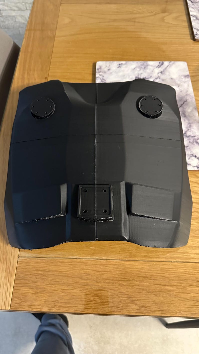 May include: A black, angular object with a symmetrical design. It has two circular components on top and a rectangular piece with four holes in the centre. The object appears to be made of a solid, possibly 3D-printed material.