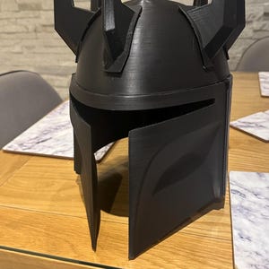 Puede incluir: Casco negro con un diseño distintivo, con visera y múltiples protuberancias angulares. El casco está hecho de un material oscuro y descansa sobre una mesa de madera. El diseño sugiere un tema futurista o de fantasía.