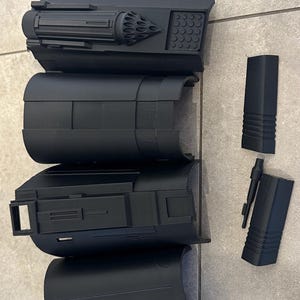 Op de afbeelding: Zwarte, 3D-geprinte componenten, waarschijnlijk voor een rekwisiet of model. De onderdelen hebben cilindrische en rechthoekige vormen, met gedetailleerde texturen en ontwerpen. De stukken zijn op een licht oppervlak geplaatst.