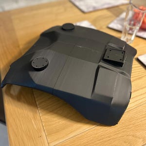 May include: A dark grey, curved component with circular and rectangular fixtures. The object, made from a matte material, sits on a wooden table. The design suggests a part of a larger structure or a component, possibly for a mechanical or technological application.