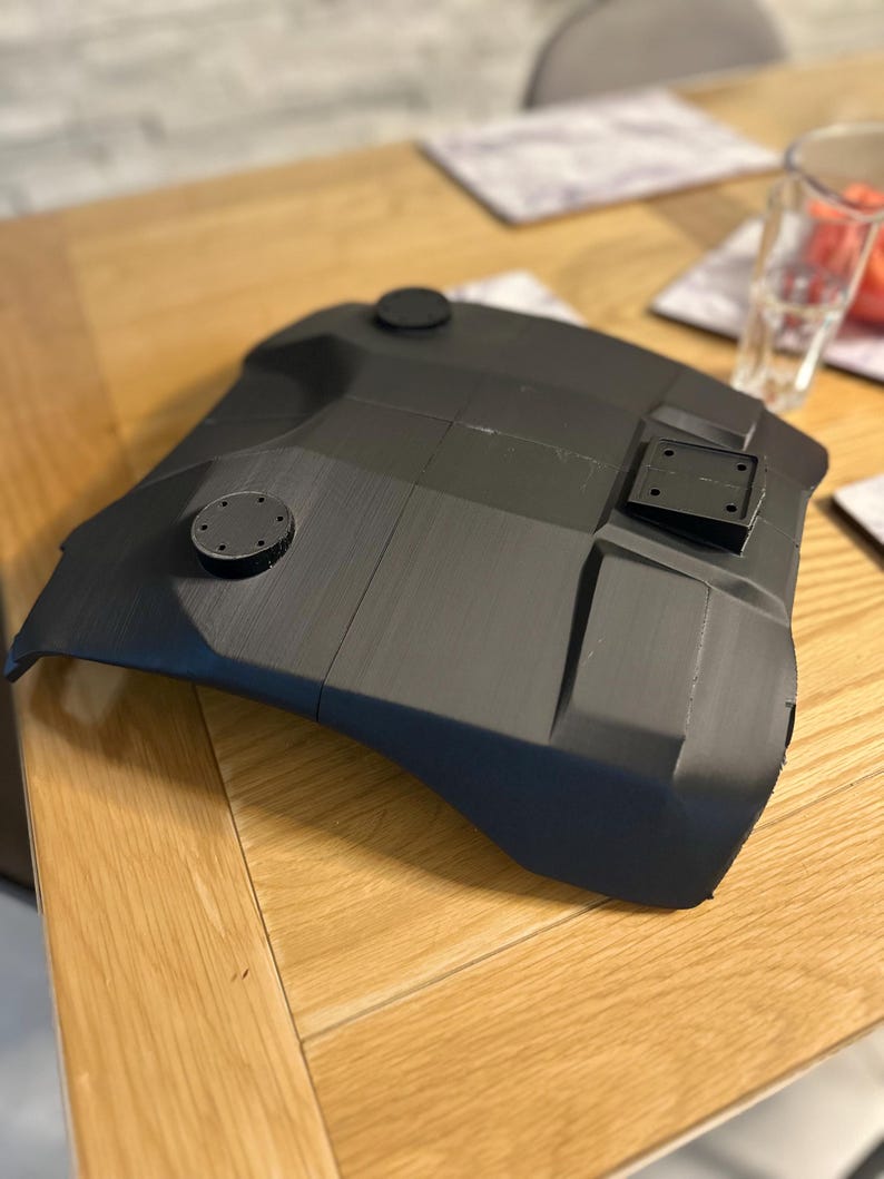 May include: A dark grey, curved component with circular and rectangular fixtures. The object, made from a matte material, sits on a wooden table. The design suggests a part of a larger structure or a component, possibly for a mechanical or technological application.