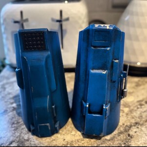 May include: Two blue, weathered cylindrical objects with rectangular details and a textured top. The objects have a slightly distressed appearance, with visible wear and tear. The background includes a white toaster.