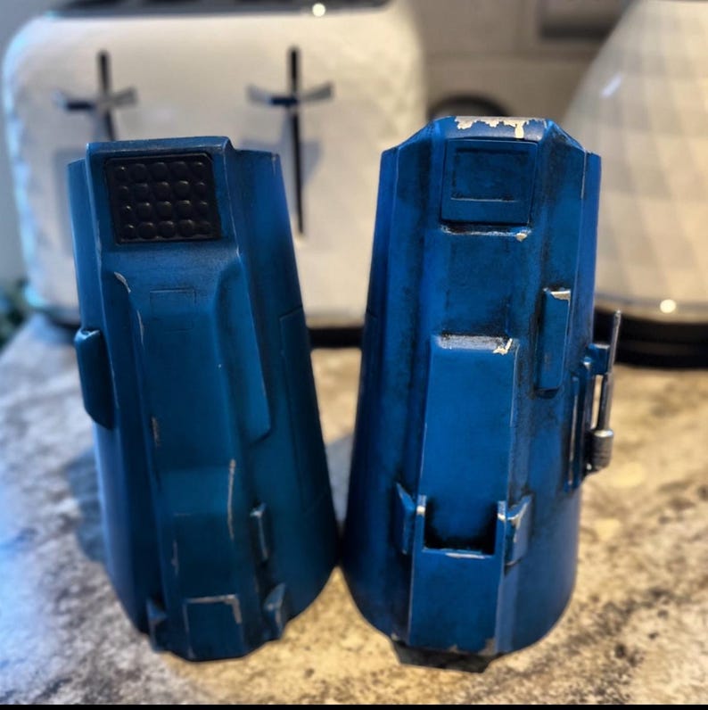 May include: Two blue, weathered cylindrical objects with rectangular details and a textured top. The objects have a slightly distressed appearance, with visible wear and tear. The background includes a white toaster.