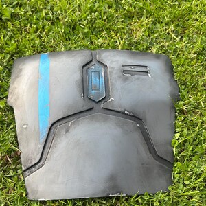 May include: A dark grey chest plate with a blue vertical stripe and a rectangular blue accent. The armour piece is made of a hard material and has a weathered appearance. The item is resting on green grass.
