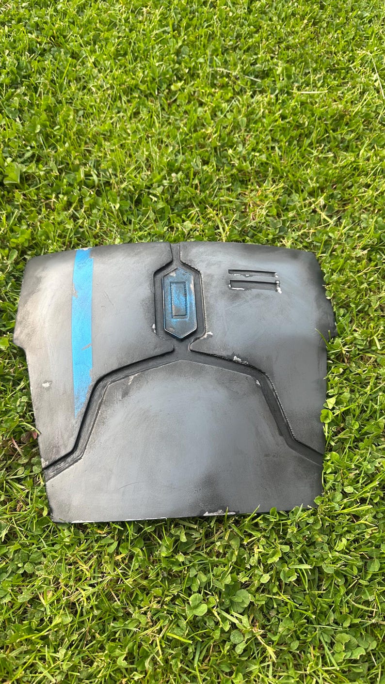 May include: A dark grey chest plate with a blue vertical stripe and a rectangular blue accent. The armour piece is made of a hard material and has a weathered appearance. The item is resting on green grass.