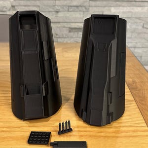May include: Black 3D-printed components, including two large, cylindrical structures and several smaller parts. The pieces are arranged on a wooden surface, suggesting a model or prototype. The design features geometric shapes and a matte finish.