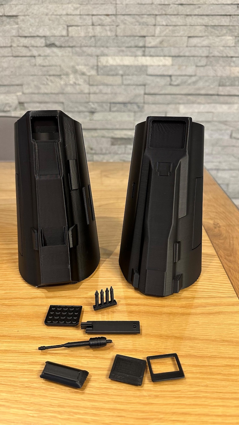 May include: Black 3D-printed components, including two large, cylindrical structures and several smaller parts. The pieces are arranged on a wooden surface, suggesting a model or prototype. The design features geometric shapes and a matte finish.