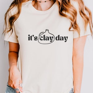 May include: A white t-shirt with a black graphic of a clay jug and the text "it's clay day".
