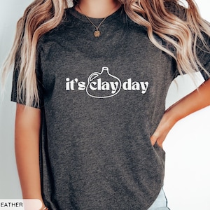 May include: A dark grey heather t-shirt with a white graphic of a clay jug and the text "it's clay day".