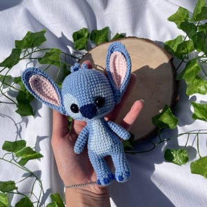 May include: A handmade blue crochet doll with large ears and black eyes. The doll has pink inner ears and a lighter blue belly. The doll is held in a hand against a backdrop of greenery and a wooden slice.