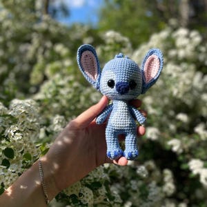May include: A handmade blue crochet doll with large ears, black eyes, and a dark blue nose. The doll has a light blue body with darker blue accents on the feet and arms. The ears have a pink interior. The doll is a character from a popular animated film.