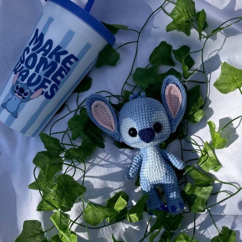 May include: A light blue crochet Stitch doll with pink inner ears and black eyes. A matching blue and white tumbler with the text "MAKE SOME WAVES" and a Stitch graphic is also present. Green vines surround the items.