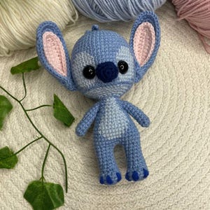 May include: A handmade blue crochet doll with large ears, black eyes, and a dark blue nose. The doll has a light blue body with darker blue accents and is standing upright. The ears have a pink interior. The doll is a character from a popular animated film.