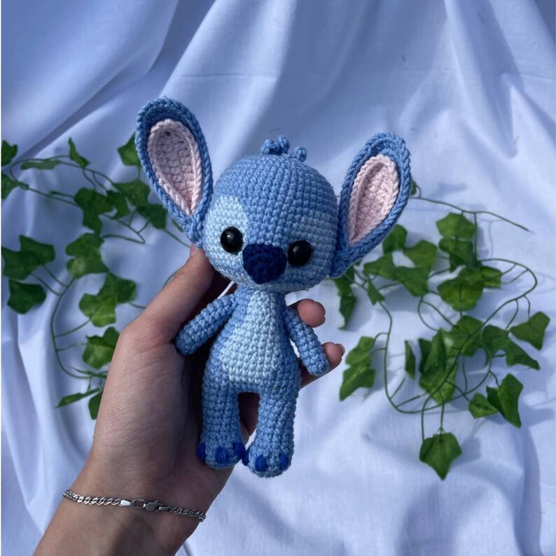 May include: A handmade blue crochet Stitch doll with large ears, black eyes, and a dark blue nose. The doll is held against a white background with green vines. The ears have a pink interior.