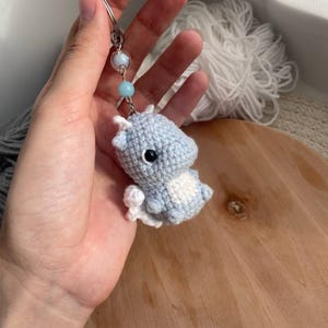 May include: A handmade crochet keychain featuring a light blue and white dragon. The dragon has black eyes and is attached to a silver keyring with blue and white beads. The dragon is approximately 2 inches tall.