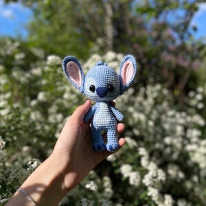 May include: A light blue crochet figure with large ears and black eyes. The figure is held in a hand against a backdrop of white flowers and green foliage. The figure's ears have pink inner details.