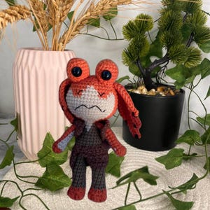Op de afbeelding: Een handgemaakte gehaakte pop met een personage met een oranje hoofd en oren, grote zwarte ogen en een wit gezicht. De pop draagt een donkergrijze en kastanjebruine outfit. Decoratieve planten en een roze vaas staan op de achtergrond.