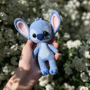May include: A handmade blue crochet doll with large ears, black eyes, and a dark blue nose. The doll has pink inner ears and feet, and is held in a hand against a blurred background of white flowers.