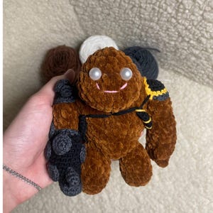 May include: A brown crocheted plush toy with a smiling face, pearl eyes, and a black and yellow shoulder detail. The toy has a dark grey arm and hand. The plush toy is held in a person's hand.