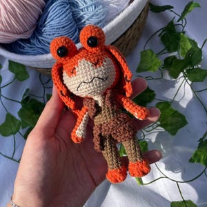 Op de afbeelding: Een handgemaakte gehaakte pop met oranje en bruin garen. De pop heeft grote zwarte ogen, oranje haar en draagt een bruin outfit. De pop wordt in een hand gehouden, met wol en groen in de achtergrond.