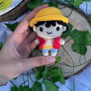 Peut inclure: Poupée au crochet faite à la main représentant un personnage portant un chapeau jaune, un gilet rouge, un pantalon bleu et une chemise jaune et bleue. La poupée a des cheveux noirs et un visage souriant. La poupée est tenue dans une main.