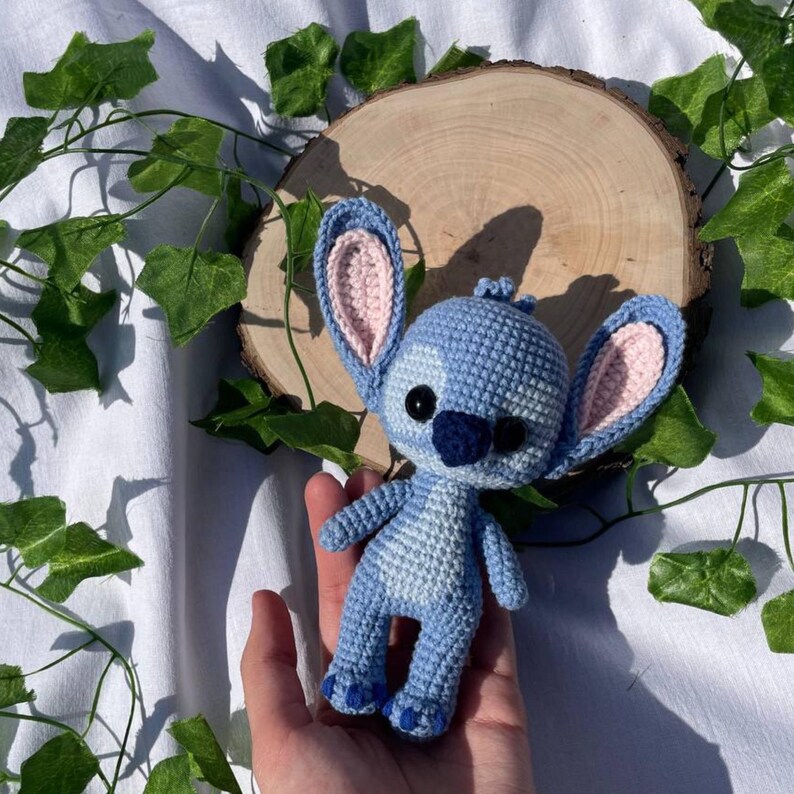 May include: A handmade blue crochet doll with large ears and a dark blue nose. The doll has pink inner ears and black eyes. The doll is held in a hand, with a wooden slice and green leaves in the background.