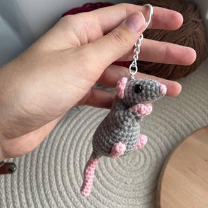 Könnte beinhalten: Handgefertigter Schlüsselanhänger in Form einer grauen Ratte mit rosa Ohren, Pfoten und Schwanz. Die Ratte hat schwarze Augen und ist an einem silberfarbenen Schlüsselring befestigt.