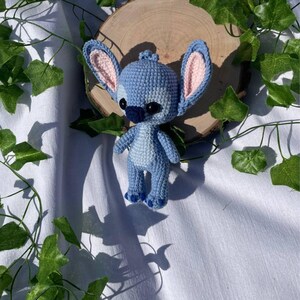 May include: A handmade crochet Stitch doll in shades of blue, with large ears and black eyes. The doll is standing on a wooden slice, surrounded by green leaves, against a white background.