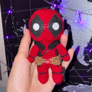 Może przedstawiać: Ręcznie robiona, czerwona szydełkowa lalka Deadpool z czarnymi akcentami wokół oczu i głowy. Lalka ma brązowy pasek i czarną kaburę. Lalka ma około 13 cm wysokości.