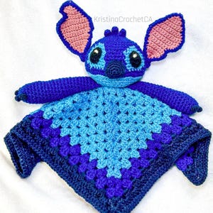 May include: A handmade crochet baby lovey featuring a blue character with large ears and a diamond-shaped blanket. The character has pink accents on the ears and a dark blue nose and eyes. The blanket transitions from light to dark blue.