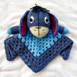 Peut inclure: Une couverture de sécurité Eeyore au crochet faite à la main. La peluche présente un corps bleu, des oreilles doublées de rose et un museau beige. La couverture est de forme carrée avec un dégradé de bleus. Le texte "KristinaCrochetCA" est visible.