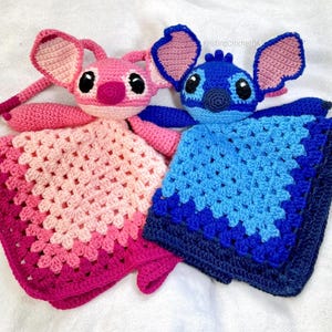 Könnte beinhalten: Zwei handgefertigte gehäkelte Baby-Kuscheltiere. Eines ist rosa mit einer passenden Decke, das andere ist blau mit einer passenden Decke. Die Kuscheltiere sind wie Zeichentrickfiguren mit großen Ohren und Augen geformt.