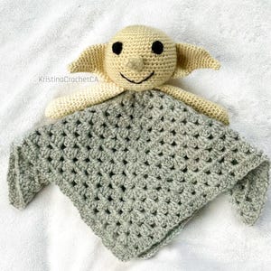 Könnte beinhalten: Eine handgefertigte Häkel-Sicherheitsdecke mit einem cremefarbenen, goblinartigen Charakter mit schwarzen Augen und einem lächelnden Mund. Der Kopf und die Arme der Figur sind an einer grauen Granny-Square-Decke befestigt. Die Decke ist aus weichem Garn gefertigt.